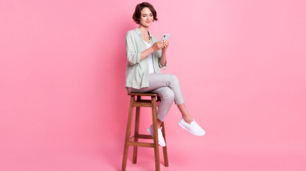 Foto em tamanho real de uma jovem sentada em um banco, usando o celular, com uma roupa cinza, isolada em um fundo rosa.