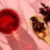 À esquerda um chá de hibisco em xícara transparente. À direita uma colher de pau com folhas de hibisco secas