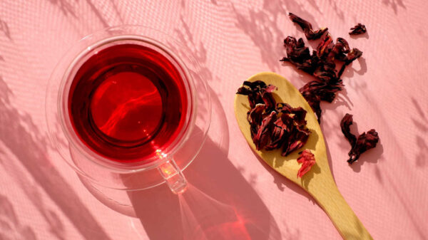À esquerda um chá de hibisco em xícara transparente. À direita uma colher de pau com folhas de hibisco secas