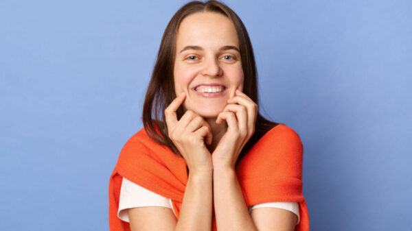 Mulher sorridente e otimista vestindo uma camiseta branca e um jumper sobre o pescoço, em pé, isolada sobre um fundo azul, de bom humor.