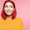 Garota de cabelo rosa, olhando de lado. Sorrindo timidamente e imaginando algo, em um fundo rosa de um estúdio. Conceito de emoções humanas.