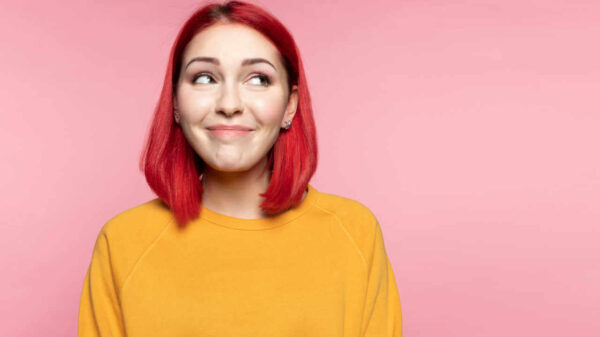 Garota de cabelo rosa, olhando de lado. Sorrindo timidamente e imaginando algo, em um fundo rosa de um estúdio. Conceito de emoções humanas.