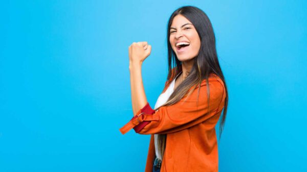 Jovem sentindo-se feliz, satisfeita e poderosa, flexionando o braço e mostrando o bíceps, contra uma parede plana.