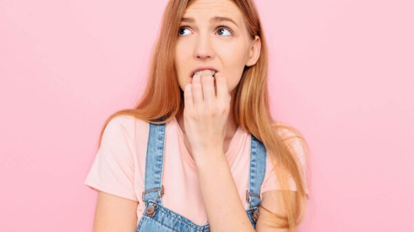 Jovem nervosa, insegura, assustada, olhando para longe, sentindo medo, insegurança e agitação, roendo as unhas, em um fundo rosa isolado.