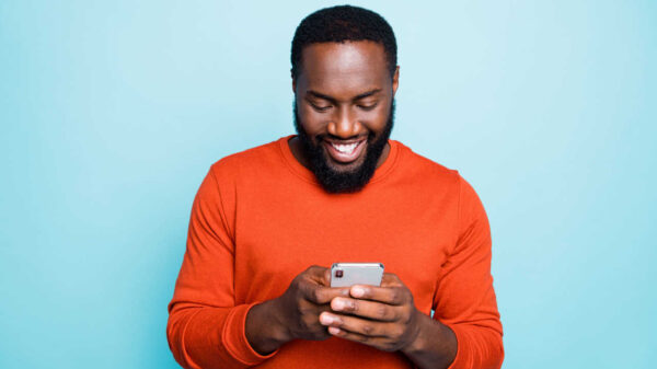 homem negro sorrindo olhando para o celular que está em suas mãos em fundo azul