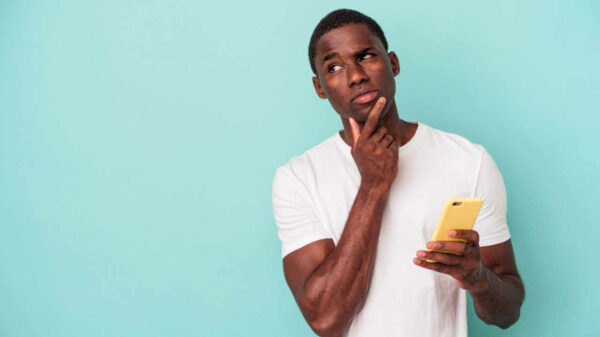 Jovem segurando um celular, isolado em um fundo azul, olhando de soslaio, com expressão de dúvida.
