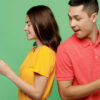 Casal de amigos jovens, (homem e mulher) usando camisetas básicas, juntos, cada um segurando um celular nas mãos, usando-os. Homem espiando o seu celular da mulher. De costas um para o outro, isolados em um fundo de cor verde pastel.