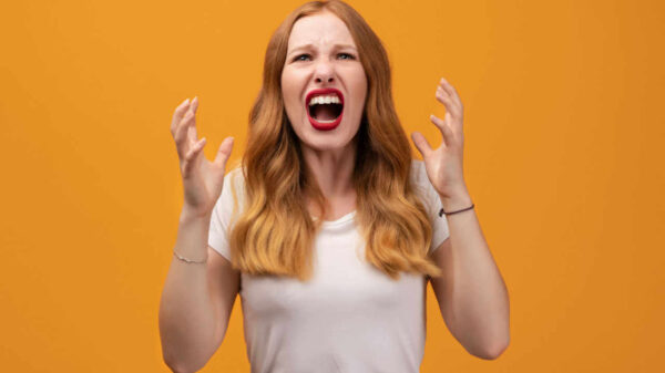 Jovem ruiva de cabelo ondulado, vestindo camiseta branca, gritando, em um fundo laranja de um estúdio. Conceito de emoções humanas e de expressão facial.