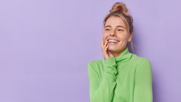 mulher branca e loira sorrindo. Ela está com a mão direita na altura no queixo. Ela veste verde. O fundo é lilás.