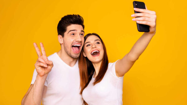 Retrato de homem e mulher alegres, positivos tirando selfie. Homem fazendo sinal de "v" com a mão. Isolados sobre um fundo amarelo.