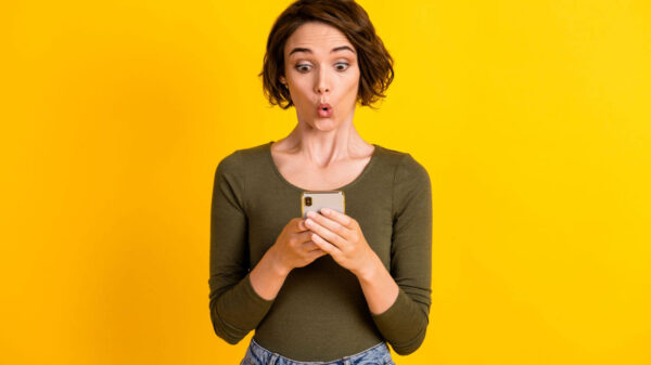 Foto de uma jovem segurando um celular, chocada, usando camisa verde, isolada em um fundo de cor amarela.