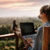 mulher jovem e loira sentada numa varanda de frente para uma paisagem com muitas árvores vista de cima. Ela tem um notebook no colo