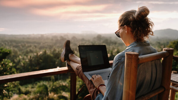 mulher jovem e loira sentada numa varanda de frente para uma paisagem com muitas árvores vista de cima. Ela tem um notebook no colo