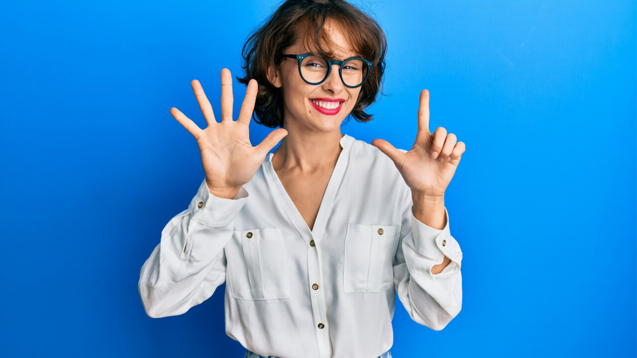 Mulher morena vestindo roupas casuais e usando óculos, fazendo com os dedos o número sete, enquanto sorri confiante e feliz.