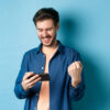 homem com expressão de felicidade segurando um celular na mão esquerda em fundo azul