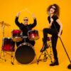 Foto de corpo inteiro de um grupo de rock. Homem tocando bateria e mulher cantando em um microfone. Conceito de banda popular. Os dois vestem roupas pretas modernas, e estão isolados em um fundo amarelo.
