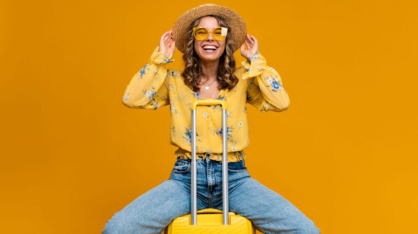 Mulher loira sorridente, feliz, viajando com uma mala, vestida com roupas modernas, usando blusa, chapéu e óculos de sol, em um fundo amarelo, Conceito de férias.