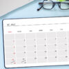 Vista superior de um notebook, de um tablet mostrando o calendário de julho de 2023, de uma caneta e de um óculos, em um fundo azul.