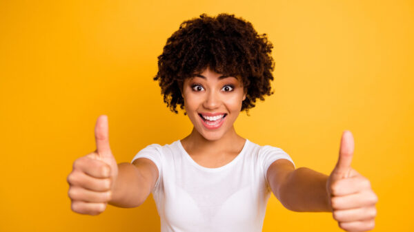 Foto de uma mulher com os braços esticados, com os dedos polegares das duas mãos para cima, vestindo camiseta branca casual, isolada em um fundo amarelo.