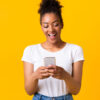 Mulher feliz, segurando um celular, sobre uma parede amarela de um estúdio.