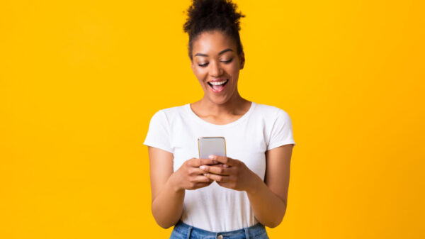 Mulher feliz, segurando um celular, sobre uma parede amarela de um estúdio.