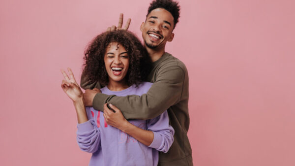 Mulher de suéter roxo piscando um olho. Homem e mulher fazendo sinal de paz em com a mão em um fundo rosa. Homem de camisa marrom. Os dois sorriem, isolados.