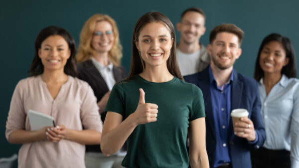 Mulher jovem feliz, com o polegar para cima, com uma equipe de pessoas diversificadas sorrindo em segundo plano.