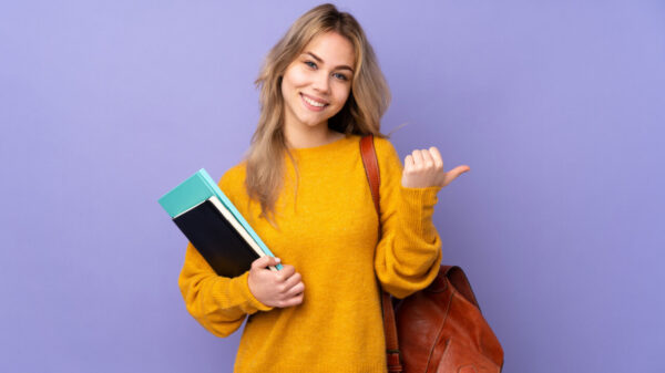 mulher loira vestindo moletom amarelo. no ombro direito está uma bolsa e na mão esquerda está cadernos