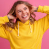 imagem de mulher branca com fios de cabelos loiro. ela veste moletom amarelo e o fundo é rosa