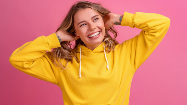 imagem de mulher branca com fios de cabelos loiro. ela veste moletom amarelo e o fundo é rosa