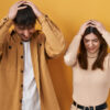 Jovem casal em pé, sobre fundo amarelo, sofrendo, desesperados e estressados, com as mãos na cabeça.