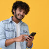 Homem sorrindo, usando um celular, isolado em um fundo amarelo.