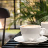 duas xícaras de café no canto inferior direito da imagem. O fundo é um local com paredes de vidro e plantas. o lugar é semelhante a uma cafeteria