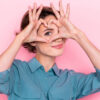 Retrato de uma mulher com os dedos fazendo o formato de um coração na frente de um de seus olhos, isolada em um fundo de cor rosa.