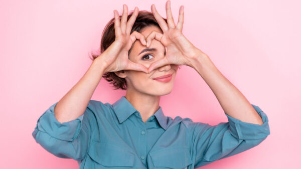 Retrato de uma mulher com os dedos fazendo o formato de um coração na frente de um de seus olhos, isolada em um fundo de cor rosa.