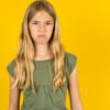 Menina ofendida e insatisfeita, vestindo uma camiseta verde, sobre um fundo amarelo, com uma expressão mal-humorada e descontente com algo.