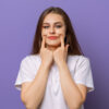 mulher branca e jovem. sua expressão facial é séria e suas mãos estão esticando a boa para formar um sorriso. o fundo é roxo