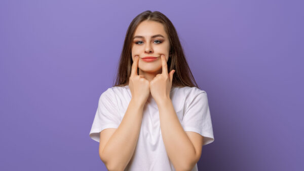 mulher branca e jovem. sua expressão facial é séria e suas mãos estão esticando a boa para formar um sorriso. o fundo é roxo