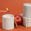 duas velas brancas e grossas. à vela que está ao centro está sendo acessa com fósforo. há duas maçãs próximas as velas. o fundo é um vermelho alaranjado