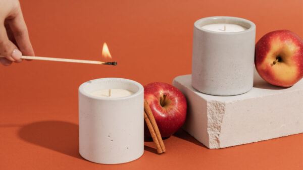 duas velas brancas e grossas. à vela que está ao centro está sendo acessa com fósforo. há duas maçãs próximas as velas. o fundo é um vermelho alaranjado