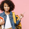 criança negra vestindo roupa branca e jaqueta jeans. no ombro esquerda está uma mochila amarela e no braço direito ela carrega três livros. o fundo é rosa