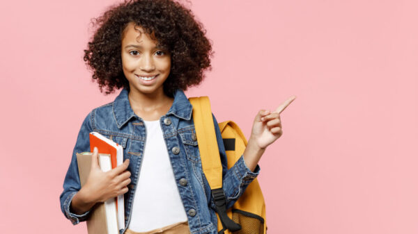 criança negra vestindo roupa branca e jaqueta jeans. no ombro esquerda está uma mochila amarela e no braço direito ela carrega três livros. o fundo é rosa