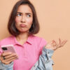 Mulher descontente, com uma expressão de frustração, segurando um celular, usando uma camiseta rosa e uma jaqueta jeans.