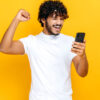 Homem surpreso e animado, segurando smartphone, recebendo notícias inesperadas, em um fundo laranja isolado, com uma expressão facial alegre, gesticulando com o punho.