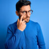 Homem com barba vestindo um suéter casual e usando um óculos, sobre um fundo azul, apontando para o próprio olho.