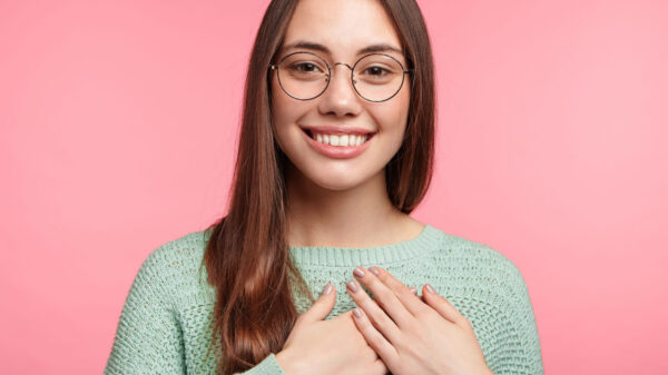 Mulher com cabelos longos, usando óculos redondos, mantendo as mãos no peito, posando contra um fundo rosa. Conceito de pessoas e emoções.