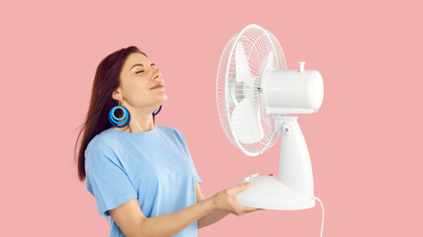 mulher jovem vestindo roupa azul e segurando nas mãos um ventilador branco em fundo rosa