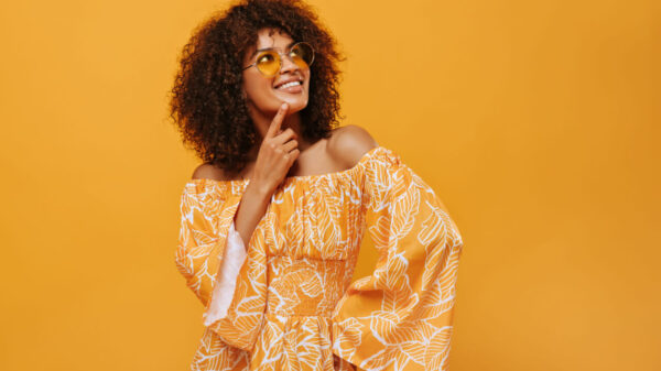 Mulher usando um óculos escuros, sorrindo, em um cenário isolado, pensativa, posando em em um fundo amarelo.
