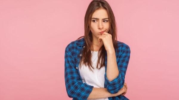 Retrato de uma mulher pensativa e chateada, usando uma camisa xadrez, ponderando sobre questões sérias, olhando para o lado com expressão hesitante e incerta. Foto de estúdio interno isolada em um fundo rosa.
