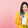 Retrato de uma mulher feliz, sorrindo, animada, surpresa, usando um celular, em um estúdio isolado em um fundo rosa.